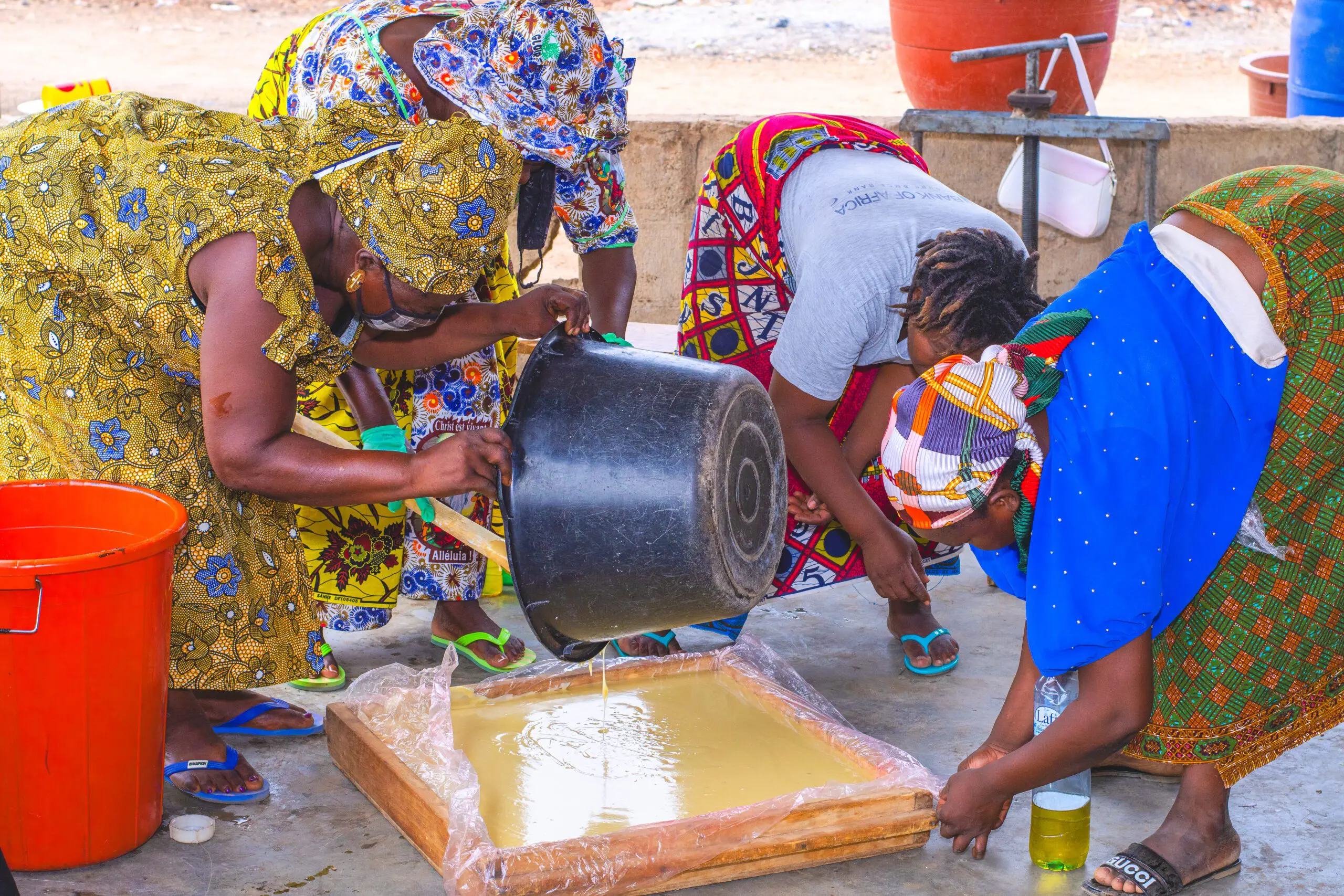 Mehrere Frauen in farbenfroher, traditioneller Kleidung beugen sich über eine Holzform, die mit Plastikfolie ausgelegt ist. Eine von ihnen giesst eine goldgelbe, flüssige Masse – frisch gewonnene, angereicherte Sheabutter – aus einem grossen schwarzen Eimer in die Form. Weitere Utensilien und Eimer stehen im Hintergrund. Die Szene zeigt einen zentralen Arbeitsschritt in der lokalen Produktion von Seife aus Sheabutter.