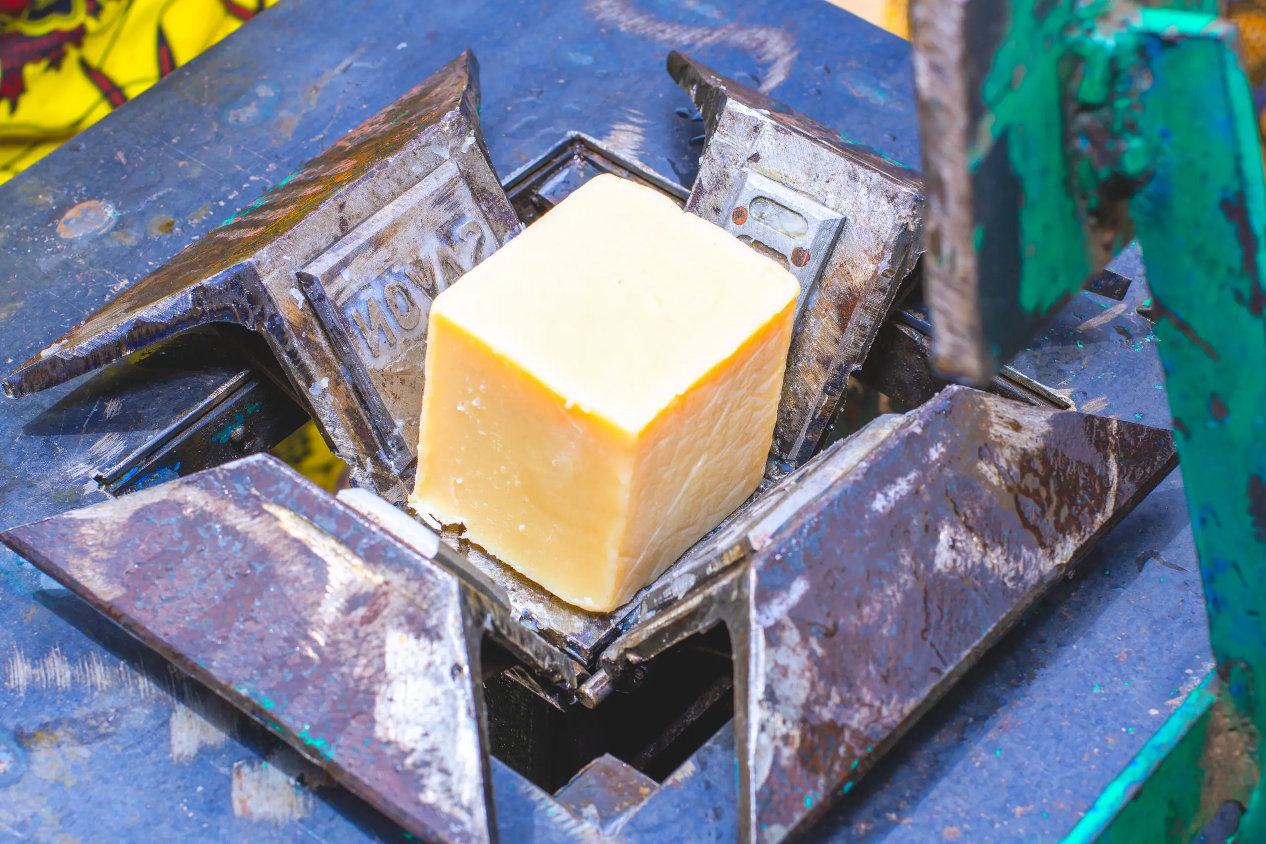 Ein frisch gepresster, heller Seifenblock aus Sheabutter liegt in einer Metallformpresse mit robustem Stahlrahmen. Die Form trägt die Prägung „SAVON“, das französische Wort für Seife. Der Block ist sauber geformt und bereit für den nächsten Verarbeitungsschritt. Das Bild zeigt einen Teil der handwerklichen Herstellung naturbelassener Seife in einer lokalen Produktionsstätte.